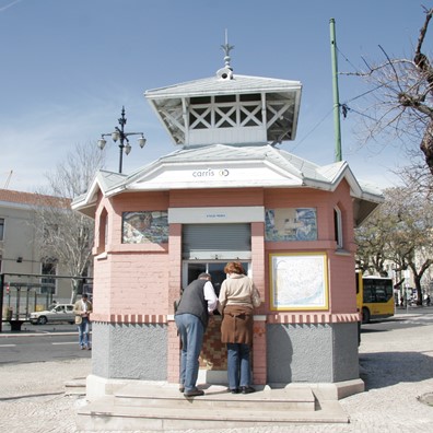 Fotografia do quiosque Carris no Cais do Sodré, com a Estação de comboios ao fundo, onde um casal está a ser atendido no guichet