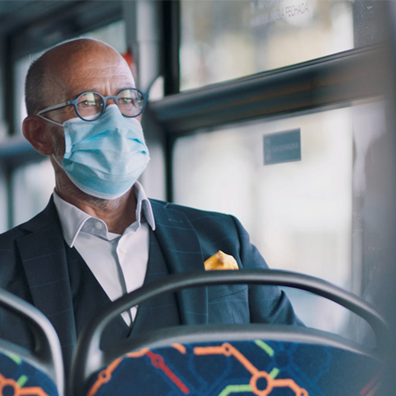 Fotografia de homem sentado em banco de autocarro, com óculos, máscara, lenço no bolso do casaco